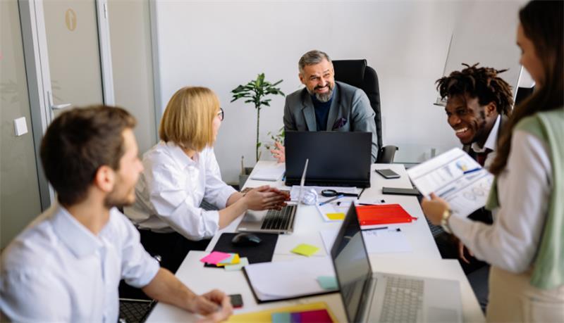Five people in business clothing at an office conference table going over documents during a meeting.