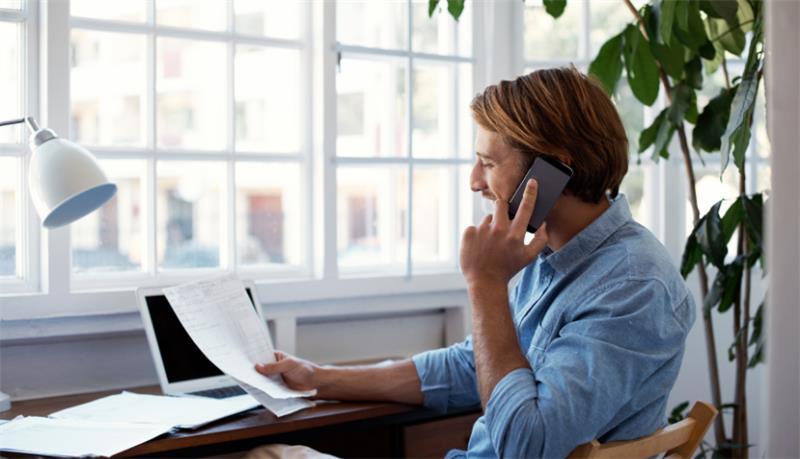A man in a blue shirt talking on a cell phone while looking at a piece of paper in his hand and sitting at a desk.