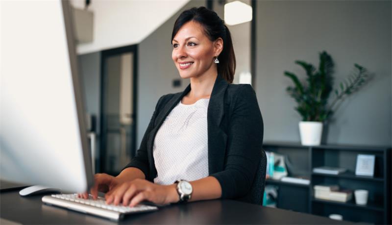 A women in a white shirt and blazer in an office smiling at a computer monitor.