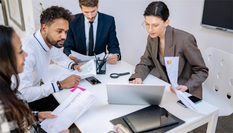 Four people in business attire sitting at a small table in an office reviewing documents. 